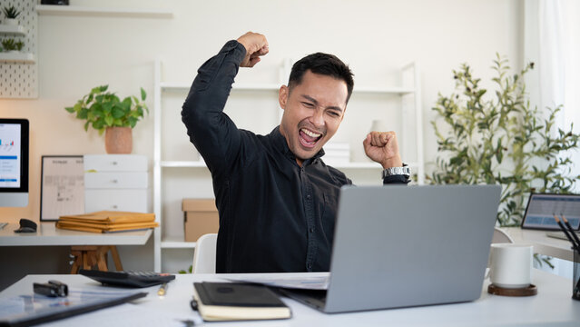 Excited businessman celebrating success while working on laptop in modern office