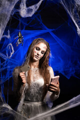 Fototapeta premium Sinister creepy woman with horrible scary Halloween zombie makeup in white dress making online shopping paying with credit card standing on blue background surrounded spiderweb. Devil person.