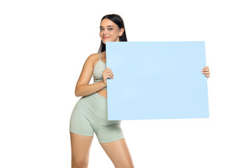 Studio shot of young woman in mint green activewear smiling and holding blank blue board for text or advertising on white background, partial studio equipment visible.