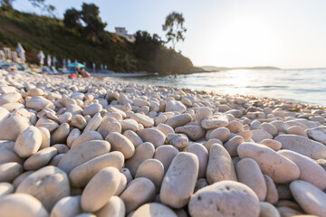 Obraz premium An up-close, low-angle shot of smooth, sun-warmed white pebbles on a beach with the bright sun creating a beautiful lens flare over the gently moving, blurred ocean.