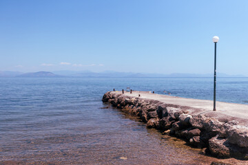 rustic stone pier stretches into the tranquil, clear blue sea under a bright sky, with hazy mountains visible on the distant horizon, evoking peace and simplicity.