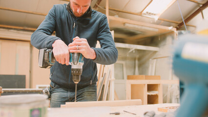 Carpenter At Workbench In Workshop Using Cordless Electric Drill On Timber