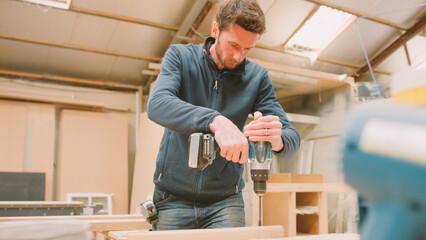 Carpenter At Workbench In Workshop Using Cordless Electric Drill On Timber