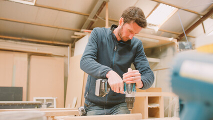 Carpenter At Workbench In Workshop Using Cordless Electric Drill On Timber