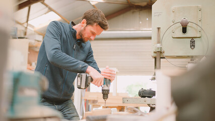 Carpenter At Workbench In Workshop Using Cordless Electric Drill On Timber