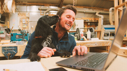 Carpenter With Pet Spaniel Working In Woodwork Workshop Using Laptop