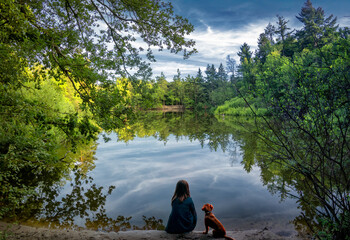 A girl and a dog sit quietly by a calm lake, surrounded by lush green trees. The water reflects the serene sky and vibrant foliage.