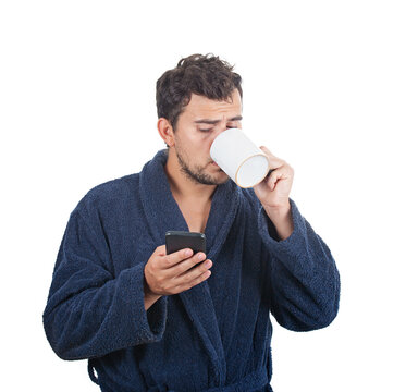 Portrait of sleepy young man messy hair, wears blue bathrobe, drinking coffee from his cup and speking on phone isolated on white background.