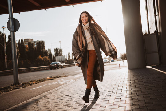 Young woman walking on an urban sidewalk in autumn, wearing a stylish coat, enjoying sunlight and expressing carefree elegance. - Powered by Adobe