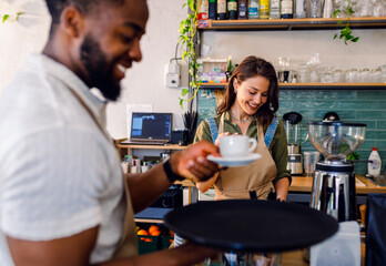 Female barista working in a coffee shop at the counter and gives a coffee to a male waiter to serve to the customer.