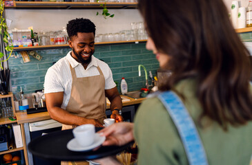 Male barista working in a coffee shop at the counter and gives a coffee to a female waiter to serve to the customer.