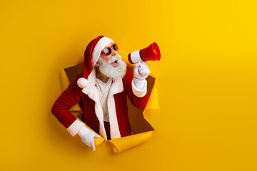 Energetic Santa Claus shouting into megaphone through yellow backdrop promoting joyful Christmas spirit and festive cheer