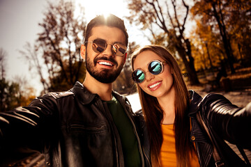 Joyful couple bonding outdoors in autumn park under sunshine, wearing stylish jackets and sunglass...
