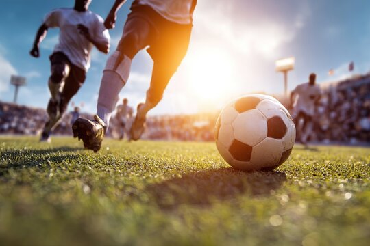 A group of diverse male soccer players running on a green field during a match. A black and white soccer ball is in the foreground with a bright sunset in the background.