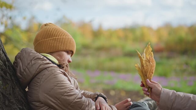 young girl rests quietly on tree trunk wearing cozy jacket and mustard beanie while mother offers bundle of dry autumn leaves and gives her a tender warm hug in colorful serene fall park setting