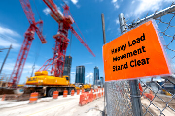A construction site featuring cranes, heavy machinery, and a safety sign warning to stand clear of heavy load movements.