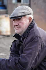 Close-up portrait of an elderly man wearing a flat cap and jacket, thoughtful expression, outdoor street style photo.