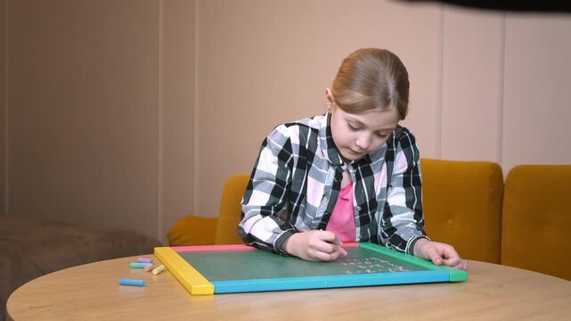 Primary school student practicing drawing on a small blackboard placed on a round wooden table near a mustard-colored sofa in a cozy living room in an apartment