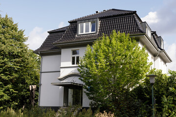 Exterior view of a large white house, featuring a dark tiled roof, surrounded by green trees and plants. Perfect for real estate or h