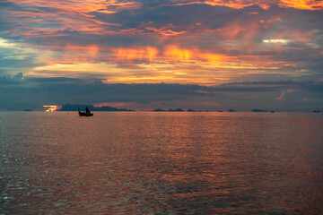 Fototapeta premium Small Boat Floating on Calm Sea During Dramatic Sunset with Fiery Sky and Island Silhouettes on Horizon, captured in wide angle, Concept Tranquil Evening Seascape