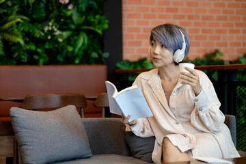 Fototapeta premium Asian adult woman reading book wearing headphones drinking coffee relaxing modern cafe environment for leisure education comfort