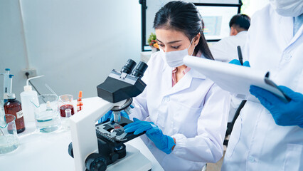 Female scientist using a microscope to examine a blood sample while a colleague records data, symbolizing teamwork, healthcare diagnostics, virus testing, and innovation in medical research.