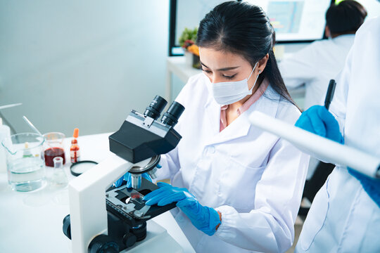 Female scientist using a microscope to examine a blood sample while a colleague records data, symbolizing teamwork, healthcare diagnostics, virus testing, and innovation in medical research.