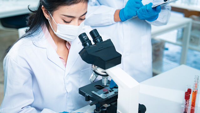 Female scientist examining a blood sample through a microscope while a colleague records notes, representing healthcare diagnostics, virus testing, laboratory research, and scientific innovation. - Powered by Adobe