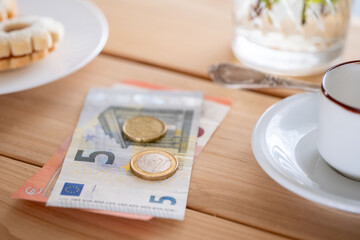 Euro banknotes and coins on wooden table with cookie and coffee cup nearby casual dining setting, Euro currency placed beside snack and coffee suggesting small purchase or tip, hospitality