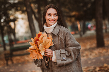 Pretty woman holding autumn leaves in a park, wearing a cozy coat and smiling joyfully, surrounded...