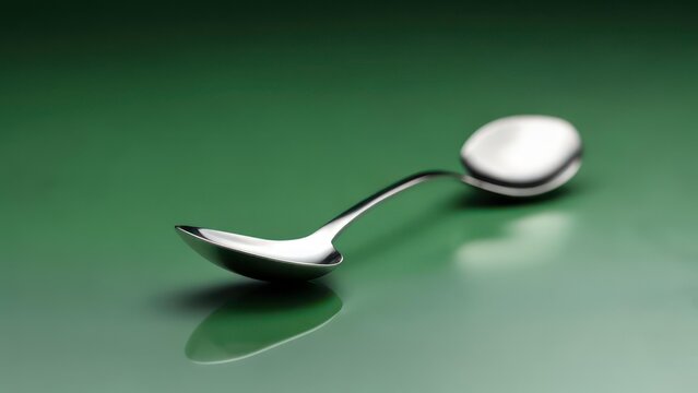 A polished silver spoon with a curved handle rests on a dark green reflective surface; its reflection is visible