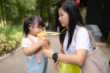Cute girl drinking water from bottle held by mom at playground ,An Asian mother feeds her daughter water from a bottle while she is walking in the park.