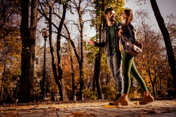 Fototapeta premium Young couple enjoying a romantic walk in an urban park surrounded by beautiful autumn foliage during sunny weather