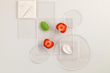 Sliced strawberries and a mint leaf are artfully placed on transparent glass geometric shapes, complemented by a swatch and a square of moisturizer texture, creating a minimalistic composition.