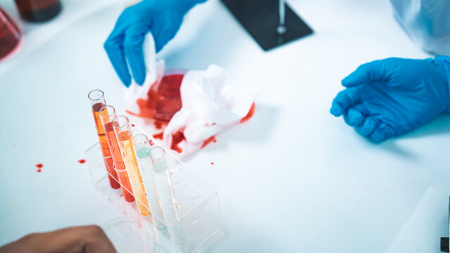 A laboratory accident shows red liquid spilled near test tubes as gloved hands attempt cleanup, symbolizing contamination risks, safety awareness, and the importance of strict lab protocols.