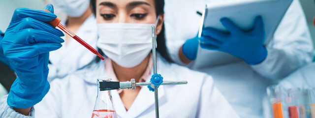 A female scientist drops a blood sample into an Erlenmeyer flask while colleagues record data,...