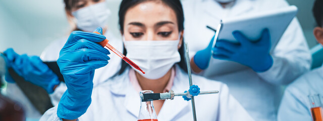 A female scientist drops a blood sample into an Erlenmeyer flask while colleagues record data,...