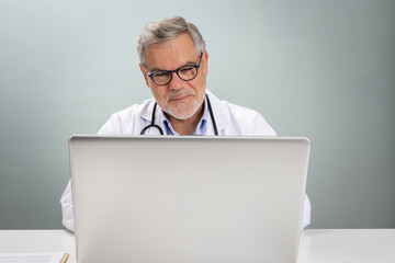 A doctor in a white coat sits at a desk, engaging in an online consultation. He appears focused while looking at a laptop screen, providing medical advice remotely.