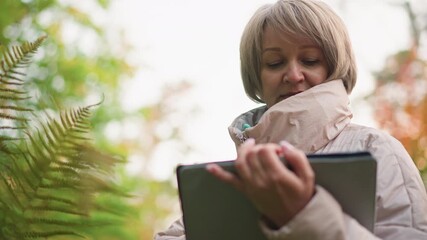 close up of female researcher observing fern leaf while recording notes on tablet in autumn forest, focused expression on face as she documents data during outdoor field study - Powered by Adobe