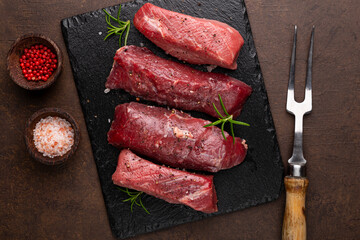 Raw beef steak. Raw fresh farm steak with rosemary, salt and pepper on black board on dark background. Raw beef steak and spices for cooking.