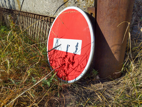 A "Do Not Enter" road sign lies on the ground, surrounded by dry grass and weeds. Its red disk is rusting, and the white bar in the center reflects shadows, creating a surreal image of refusal and obl