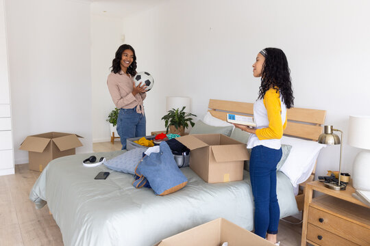 Diverse mom and teenage daughter unpacking moving boxes around bed in bedroom holding soccer ball