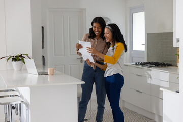 Diverse mom and teenage daughter reviewing printed documents in kitchen with laptop