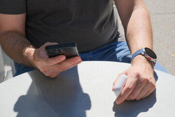 Close-up of a man at an outdoor table, holding a smartphone and earbuds case, with a smartwatch on his wrist.