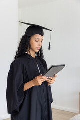 Diverse teenager standing in cap and gown holding tablet in empty studio