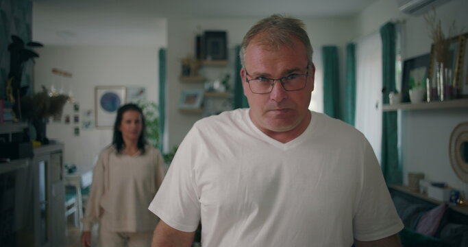 Man staring at camera with intense expression during argument, woman in background in living room, tense and emotional moment of relationship conflict and frustration
