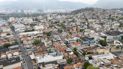Fototapeta premium Imagem aérea de área residencial próxima ao Estádio São Januário, no Rio de Janeiro, com ruas, casas, galpões, prédios e morros ao fundo sob céu cinzento e atmosfera urbana.