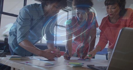 Collaborating team writing sticky notes on table near laptop at modern office, with digital overlay