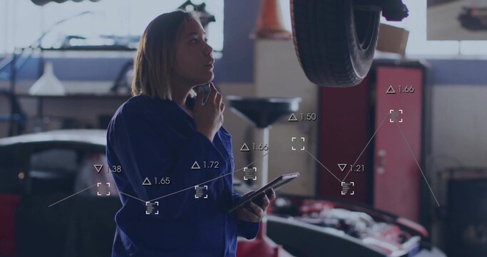 Female auto technician in coverall holding tablet while speaking on smartphone in garage, with tire - Powered by Adobe