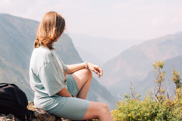 Young woman in sportswear sits on a rock ledge, peacefully observing scenic mountain landscape during a summer hiking adventure.